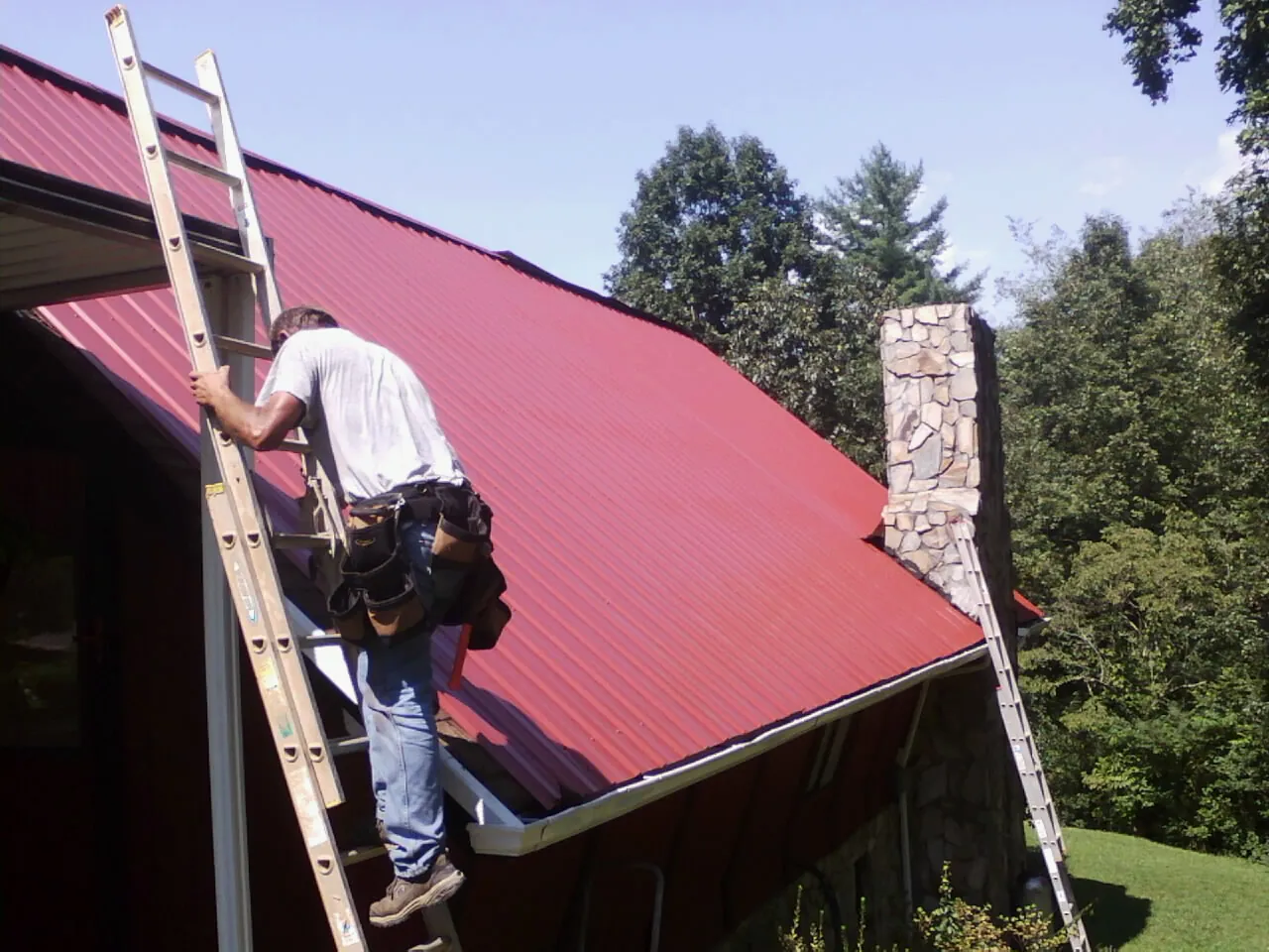 Roofer on a ladder beside a red metal roof and stone chimney — Soffit & Fascia Repair by Roofing Brothers in Boothbay Harbor